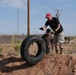 Two individuals are working together to move or position a large truck tire in a desert/arid environment. A man wearing a red cap, black t-shirt, and cargo shorts is guiding the tire while a younger person assists from behind. The setting appears to be a community cleanup or construction project in a dry, scrubland area.