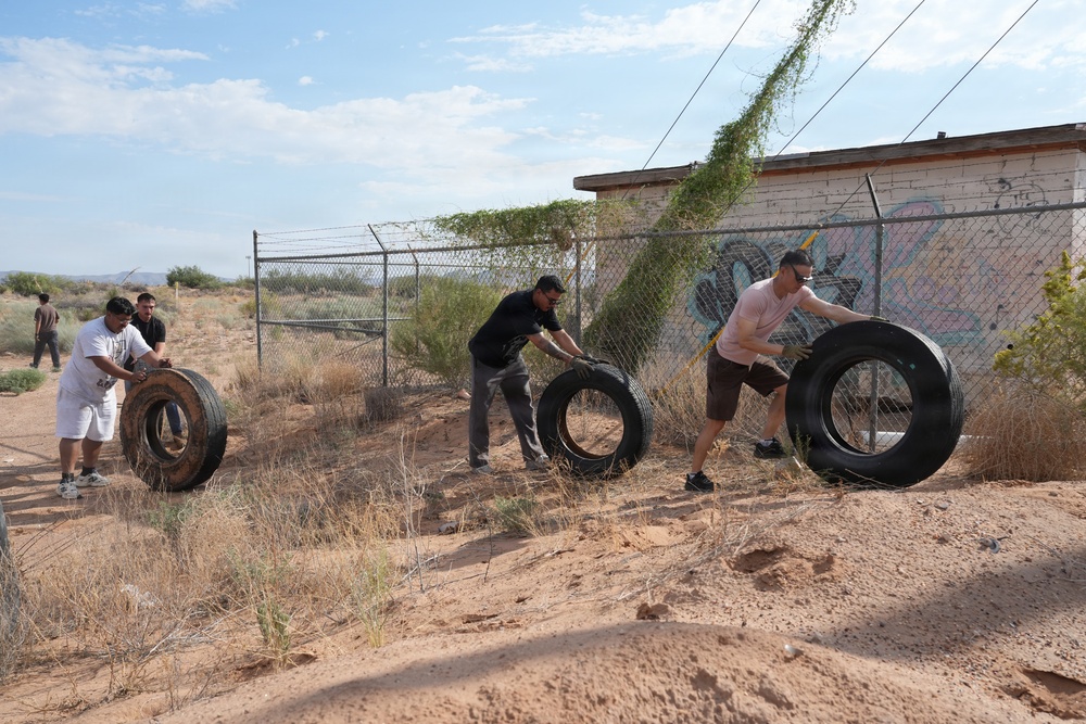 Several people are moving large tires in a desert/arid environment along a chain-link fence next to an abandoned or deteriorated building with graffiti. The activity appears to be either a cleanup effort or a physical fitness exercise. The setting is dry and sandy with sparse desert vegetation.