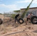 Several people are moving large tires in a desert/arid environment along a chain-link fence next to an abandoned or deteriorated building with graffiti. The activity appears to be either a cleanup effort or a physical fitness exercise. The setting is dry and sandy with sparse desert vegetation.