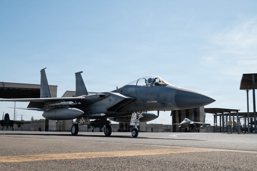A U.S. Air Force F-15 Eagle fighter jet is taxiing on a flight line apron at a military air base. The aircraft is armed with visible air-to-air missiles and carries external fuel tanks. Additional aircraft and military hangars are visible in the background.