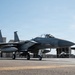 A U.S. Air Force F-15 Eagle fighter jet is taxiing on a flight line apron at a military air base. The aircraft is armed with visible air-to-air missiles and carries external fuel tanks. Additional aircraft and military hangars are visible in the background.