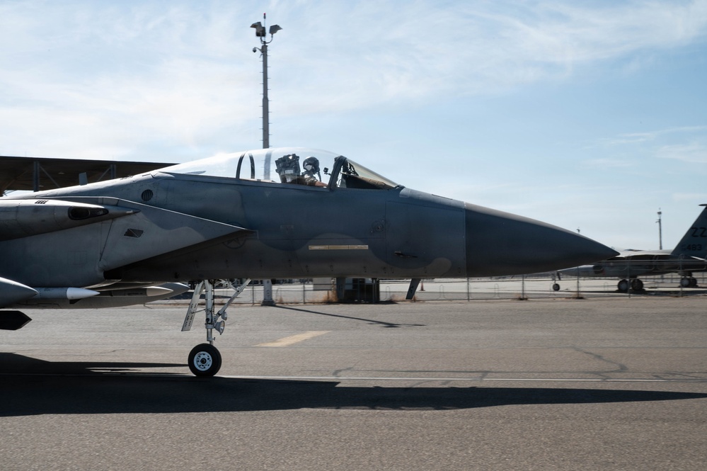 A military fighter jet, identifiable as an F-15 Eagle, is taxiing on an airfield tarmac with a pilot visible in the cockpit wearing a flight helmet and oxygen mask. The aircraft displays a two-tone gray camouflage paint scheme and has its nose gear extended. Additional aircraft and base infrastructure are visible in the background.