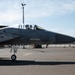 A military fighter jet, identifiable as an F-15 Eagle, is taxiing on an airfield tarmac with a pilot visible in the cockpit wearing a flight helmet and oxygen mask. The aircraft displays a two-tone gray camouflage paint scheme and has its nose gear extended. Additional aircraft and base infrastructure are visible in the background.