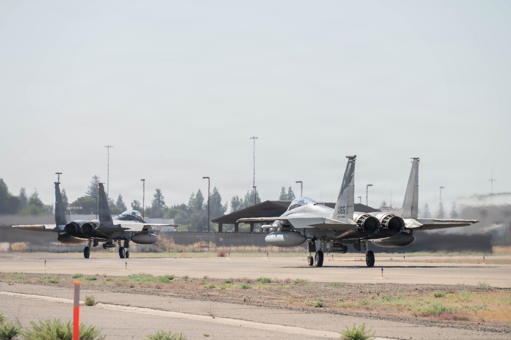 Two F-15 Eagle fighter jets are taxiing on a military airfield runway, positioned side by side. The aircraft appear to be in gray camouflage livery with engine exhaust heat distortion visible behind them. The setting appears to be a U.S. Air Force or Air National Guard base with trees, light poles, and a hangar structure visible in the background.