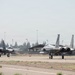 Two F-15 Eagle fighter jets are taxiing on a military airfield runway, positioned side by side. The aircraft appear to be in gray camouflage livery with engine exhaust heat distortion visible behind them. The setting appears to be a U.S. Air Force or Air National Guard base with trees, light poles, and a hangar structure visible in the background.