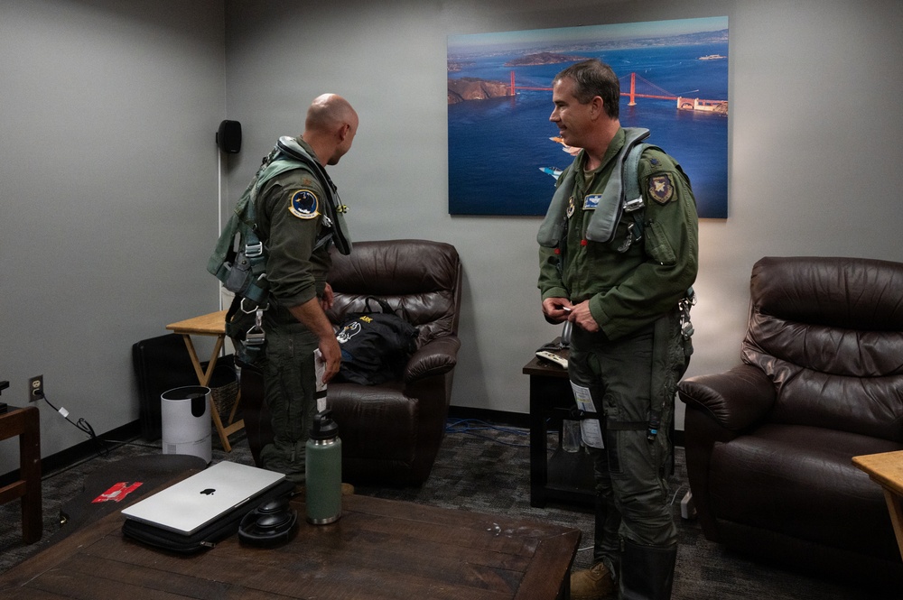 Two military aviators in olive green flight suits and survival vests are conversing in what appears to be a pilot ready room or lounge area. Both are fully equipped with flight gear including harnesses and G-suit attachments. The room contains casual furniture and a large aerial photograph of the Golden Gate Bridge on the wall.