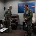 Two military aviators in olive green flight suits and survival vests are conversing in what appears to be a pilot ready room or lounge area. Both are fully equipped with flight gear including harnesses and G-suit attachments. The room contains casual furniture and a large aerial photograph of the Golden Gate Bridge on the wall.