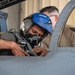 A military pilot wearing a blue helmet with the callsign 'SHARK' and an oxygen mask is seated in a fighter jet cockpit, leaning forward to inspect or adjust equipment. A second individual, wearing safety glasses, is visible behind the pilot, likely a crew chief or ground technician assisting with pre-flight preparations. The image is taken close-up near the cockpit of what appears to be a modern military jet aircraft.