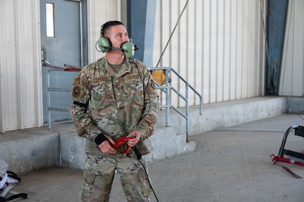 A U.S. Air Force service member named Sandoval is standing near a hangar or maintenance facility wearing OCP (Operational Camouflage Pattern) uniform. He is equipped with green aviation-style communication headset and is holding a red-handled device with a cable, likely a ground interphone or aircraft communication connector. He appears to be looking upward, possibly monitoring aircraft or awaiting instructions.