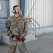 A U.S. Air Force service member named Sandoval is standing near a hangar or maintenance facility wearing OCP (Operational Camouflage Pattern) uniform. He is equipped with green aviation-style communication headset and is holding a red-handled device with a cable, likely a ground interphone or aircraft communication connector. He appears to be looking upward, possibly monitoring aircraft or awaiting instructions.