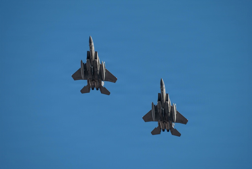 Two military fighter jets, identifiable as F-15 Eagles, are photographed from below against a clear blue sky. Both aircraft are flying in a close formation and are viewed from their ventral (underside) perspective. Multiple munitions or external stores are visible mounted beneath each aircraft.