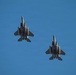 Two military fighter jets, identifiable as F-15 Eagles, are photographed from below against a clear blue sky. Both aircraft are flying in a close formation and are viewed from their ventral (underside) perspective. Multiple munitions or external stores are visible mounted beneath each aircraft.