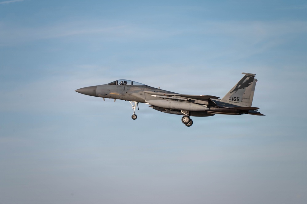 A U.S. Air Force F-15 Eagle fighter jet is captured in flight with landing gear fully extended, indicating an approach or landing phase. The aircraft bears tail markings 'AF 86-165' and the word 'CALIFORNIA,' identifying it as belonging to the California Air National Guard. The image is taken against a clear blue sky with minimal cloud cover.