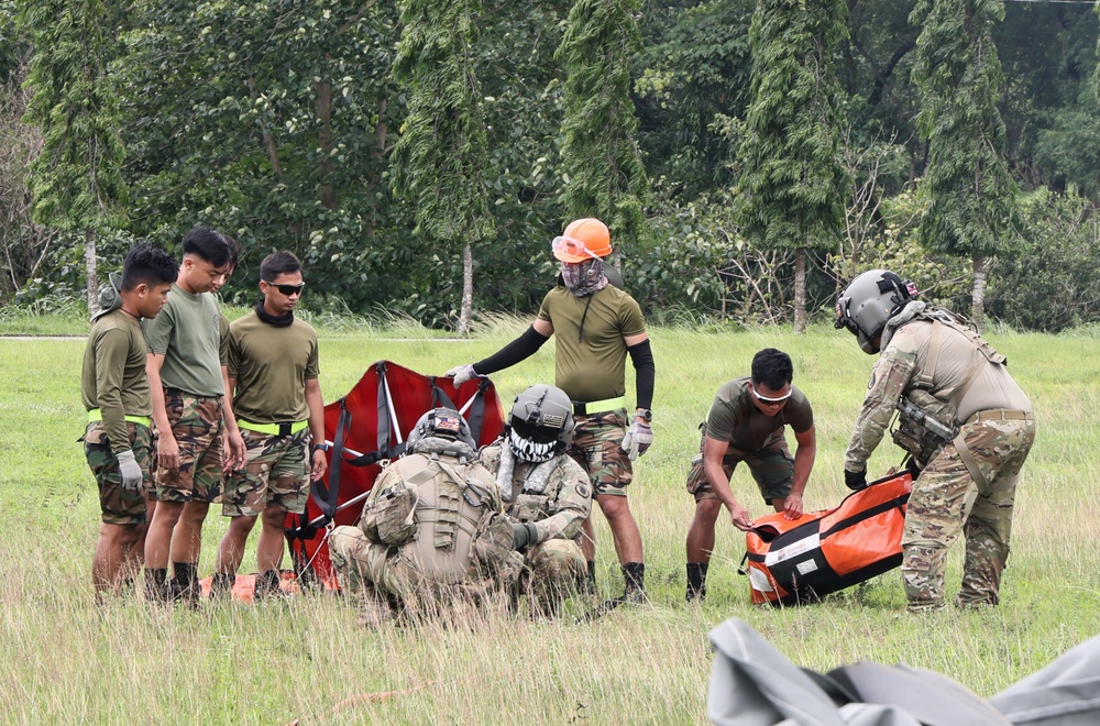 A group of military or paramilitary personnel are gathered in an open grassy field conducting what appears to be a training exercise involving rescue or extraction equipment. The group includes individuals in camouflage uniforms, tactical gear, and helmets, working together around a red litter/stretcher and an orange equipment bag. The setting appears to be a tropical or subtropical environment based on the dense green vegetation in the background.