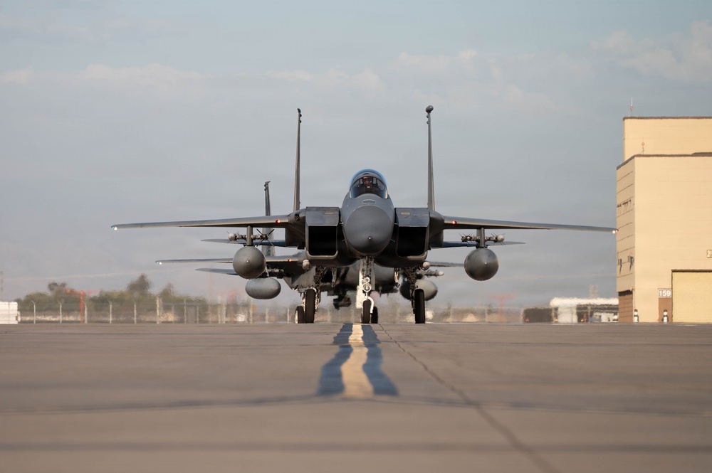 A military fighter jet, consistent with an F-15 Eagle, is taxiing directly toward the camera on an airfield tarmac. The aircraft is viewed head-on, revealing its twin-tail configuration, loaded weapons pylons, and external fuel tanks. The setting appears to be a military air base with a hangar building visible on the right side.