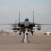 A military fighter jet, consistent with an F-15 Eagle, is taxiing directly toward the camera on an airfield tarmac. The aircraft is viewed head-on, revealing its twin-tail configuration, loaded weapons pylons, and external fuel tanks. The setting appears to be a military air base with a hangar building visible on the right side.
