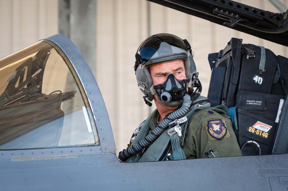 A military fighter pilot is seated in the cockpit of a jet aircraft, wearing full flight gear including a helmet with visor and an oxygen mask. The aircraft appears to be an F-16 or similar single-seat fighter jet, with visible ejection seat markings and serial number 86-0146. The image is a close-up portrait-style photograph taken on what appears to be a flight line or tarmac.