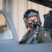 A military fighter pilot is seated in the cockpit of a jet aircraft, wearing full flight gear including a helmet with visor and an oxygen mask. The aircraft appears to be an F-16 or similar single-seat fighter jet, with visible ejection seat markings and serial number 86-0146. The image is a close-up portrait-style photograph taken on what appears to be a flight line or tarmac.