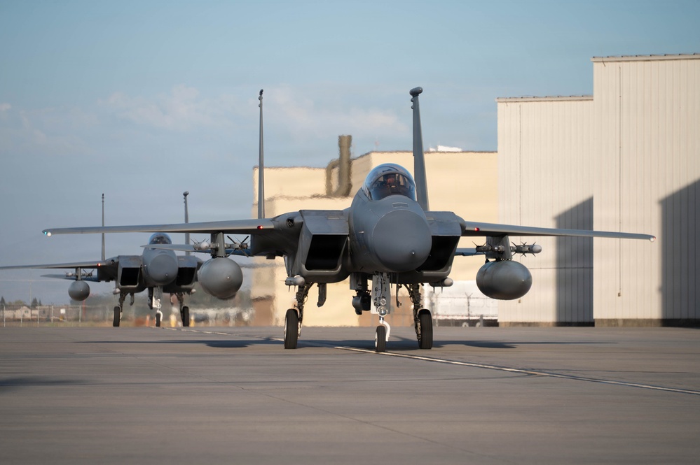 Three military fighter jets, identified as F-15 Eagles, are taxiing in formation on an airfield tarmac. The lead aircraft is prominently featured in the foreground with a pilot visible in the cockpit, while two additional aircraft follow behind. Large hangar structures are visible in the background under a partly cloudy sky.