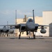 Three military fighter jets, identified as F-15 Eagles, are taxiing in formation on an airfield tarmac. The lead aircraft is prominently featured in the foreground with a pilot visible in the cockpit, while two additional aircraft follow behind. Large hangar structures are visible in the background under a partly cloudy sky.