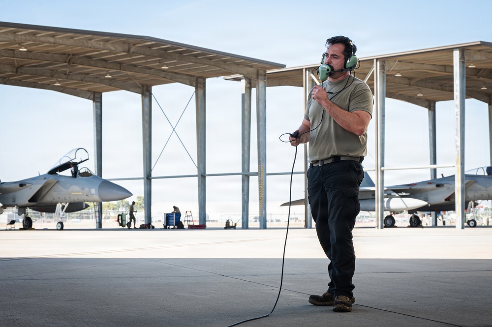 A military aircraft maintainer or ground crew member stands on a flight line apron wearing green aviation headset and holding a communication cord, likely conducting pre-flight or maintenance communications. Behind him, at least two military jet aircraft — appearing to be F-15 variants — are parked under a large open-sided canopy structure. Additional ground personnel are visible working in the background.
