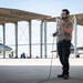 A military aircraft maintainer or ground crew member stands on a flight line apron wearing green aviation headset and holding a communication cord, likely conducting pre-flight or maintenance communications. Behind him, at least two military jet aircraft — appearing to be F-15 variants — are parked under a large open-sided canopy structure. Additional ground personnel are visible working in the background.