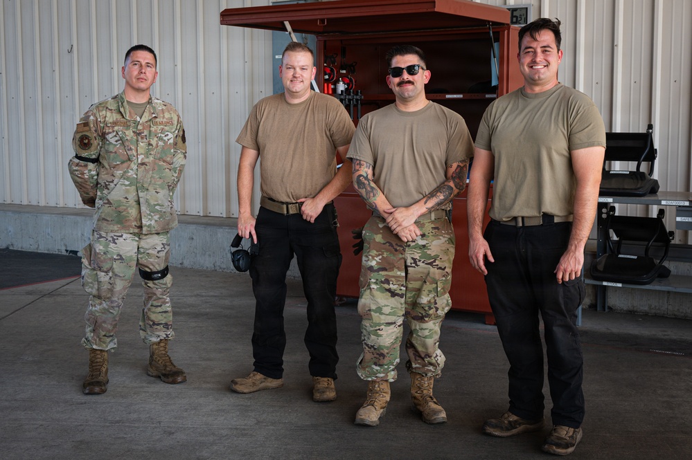 Four military personnel pose for a group photo inside what appears to be a hangar or maintenance facility. One individual on the left wears full OCP (Operational Camouflage Pattern) uniform with patches visible, while the others wear tan/coyote brown t-shirts with a mix of military and civilian-style pants. The setting suggests a maintenance or support unit environment.