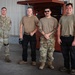Four military personnel pose for a group photo inside what appears to be a hangar or maintenance facility. One individual on the left wears full OCP (Operational Camouflage Pattern) uniform with patches visible, while the others wear tan/coyote brown t-shirts with a mix of military and civilian-style pants. The setting suggests a maintenance or support unit environment.