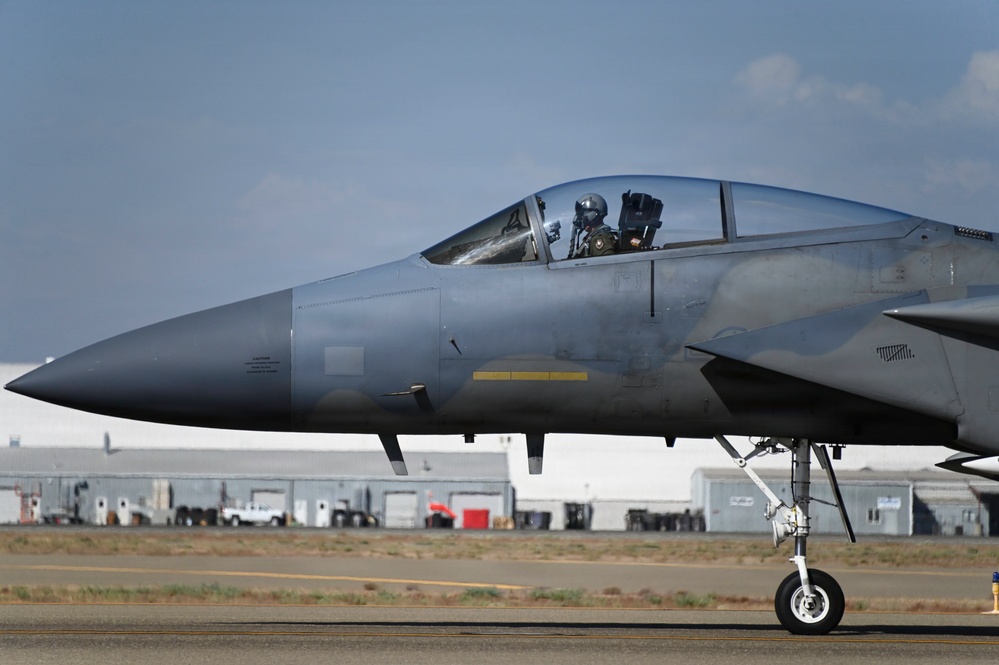 A military jet fighter, consistent with an F-15 Eagle, is taxiing on a runway at what appears to be a military air base. A helmeted pilot is visible in the cockpit with the canopy open. The aircraft is painted in a gray camouflage scheme and military support buildings are visible in the background.