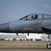 A military jet fighter, consistent with an F-15 Eagle, is taxiing on a runway at what appears to be a military air base. A helmeted pilot is visible in the cockpit with the canopy open. The aircraft is painted in a gray camouflage scheme and military support buildings are visible in the background.