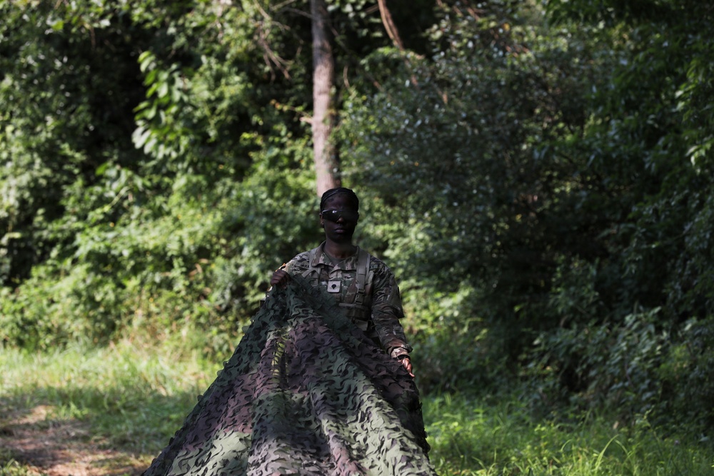 A soldier in military camouflage uniform is carrying or deploying a large camouflage netting in a forested outdoor environment. The individual has face paint applied and appears to be engaged in a field training or tactical exercise. The lush green vegetation in the background suggests a tropical or subtropical setting.