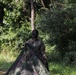 A soldier in military camouflage uniform is carrying or deploying a large camouflage netting in a forested outdoor environment. The individual has face paint applied and appears to be engaged in a field training or tactical exercise. The lush green vegetation in the background suggests a tropical or subtropical setting.