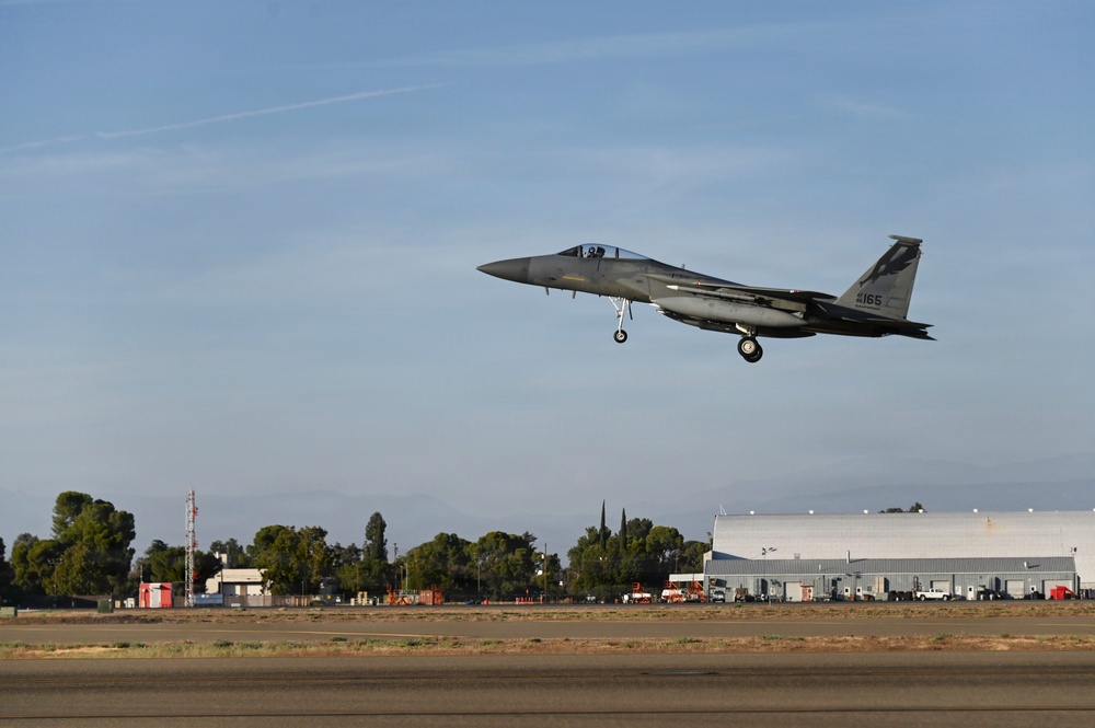 An F-15 Eagle fighter jet with tail number 165 is captured in low-altitude flight with landing gear deployed, suggesting it is on final approach for landing. The aircraft is photographed over a military airfield with hangars, support buildings, and ground vehicles visible in the background. The image was taken during daylight hours under partly cloudy skies with warm, golden-toned lighting suggesting early morning or late afternoon.