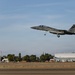 An F-15 Eagle fighter jet with tail number 165 is captured in low-altitude flight with landing gear deployed, suggesting it is on final approach for landing. The aircraft is photographed over a military airfield with hangars, support buildings, and ground vehicles visible in the background. The image was taken during daylight hours under partly cloudy skies with warm, golden-toned lighting suggesting early morning or late afternoon.