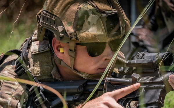 A soldier in full multicam camouflage gear is lying prone in grass, aiming a rifle with an optic sight. The individual is equipped with tactical helmet, ballistic sunglasses, ear protection, and body armor with attached gear. The image appears to depict a military training exercise or field operation.