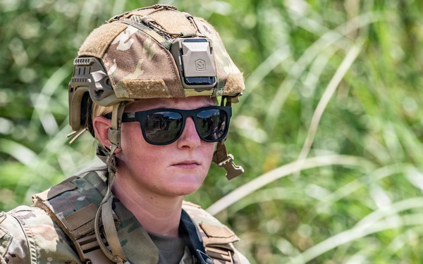 A military service member wearing MultiCam (OCP) camouflage uniform, tactical helmet, and sunglasses is photographed in an outdoor environment with dense green vegetation in the background. The individual's name tape reads 'MORTON' and they are carrying a weapon while appearing alert and scanning their surroundings. The image appears to be a professional military documentation or press photograph taken in a field training or operational environment.