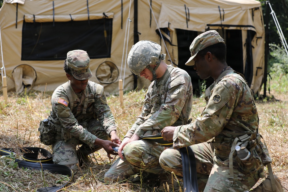 Three U.S. Army soldiers in camouflage uniforms are crouched on the ground working with cables or wiring equipment in a field setting. They appear to be conducting a field training exercise near a tan military tent structure. The soldiers are collaboratively handling what appears to be communications or electrical equipment.