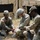 Three U.S. Army soldiers in camouflage uniforms are crouched on the ground working with cables or wiring equipment in a field setting. They appear to be conducting a field training exercise near a tan military tent structure. The soldiers are collaboratively handling what appears to be communications or electrical equipment.