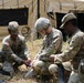 Three U.S. Army soldiers in camouflage uniforms are crouched on the ground working with cables or wiring equipment in a field setting. They appear to be conducting a field training exercise near a tan military tent structure. The soldiers are collaboratively handling what appears to be communications or electrical equipment.