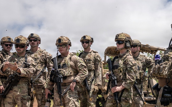 A group of approximately nine military personnel in full tactical gear are assembled outdoors in a field setting. They are wearing matching multicam camouflage uniforms, tactical helmets, and sunglasses, and are carrying rifles. A tan military utility vehicle is visible in the background, suggesting a training exercise or operational briefing.