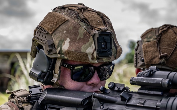 A military operator in full tactical gear is aiming a rifle around a concrete corner or wall. The soldier is equipped with modern special operations equipment including a ballistic helmet, communications gear, and optical sights. A second similarly equipped operator is visible in the background.