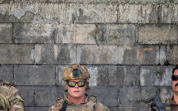 Three armed military personnel in MultiCam/OCP camouflage uniforms stand in front of a weathered concrete block wall. All three are equipped with tactical gear, body armor, and assault rifles, and are wearing dark sunglasses. The leftmost individual has a visible name tape reading 'SEXTON'; the center individual is wearing a ballistic helmet.