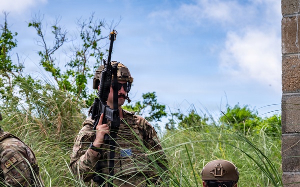 Three military personnel in full tactical gear are moving through tall grass near a concrete block structure. The soldiers are wearing camouflage uniforms, helmets, and sunglasses, with at least one visibly raising a rifle. The setting appears to be an outdoor training or operational environment with dense vegetation and a partially ruined or abandoned building.