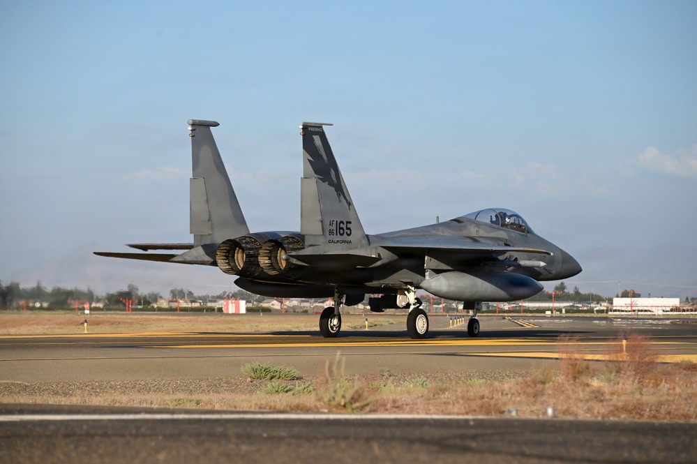 An F-15 Eagle fighter jet is taxiing on a runway at what appears to be a California Air National Guard base. The aircraft bears tail number 165 with 'AF 86' and 'CALIFORNIA' markings, and features a distinctive bird/eagle emblem on the vertical stabilizer. The location appears to be Fresno Air National Guard Base based on visible markings.