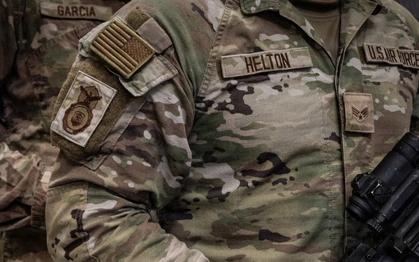 Close-up photograph of U.S. Air Force personnel in OCP (Operational Camouflage Pattern) uniforms. The central figure, identified by name tape as 'HELTON,' holds a military rifle and wears a tactical watch. A second service member identified as 'GARCIA' is partially visible in the background.