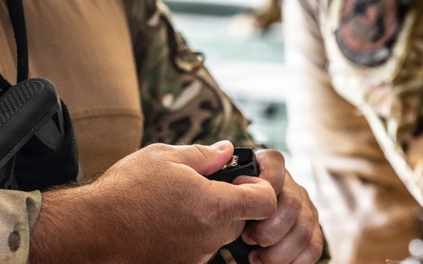 A close-up image showing the hands of a person in military camouflage uniform handling what appears to be a firearm magazine or small tactical device. The individual is wearing MultiCam/OCP pattern camouflage and has additional tactical gear visible including a holster and radio/communication device. A second person in camouflage is partially visible in the background.