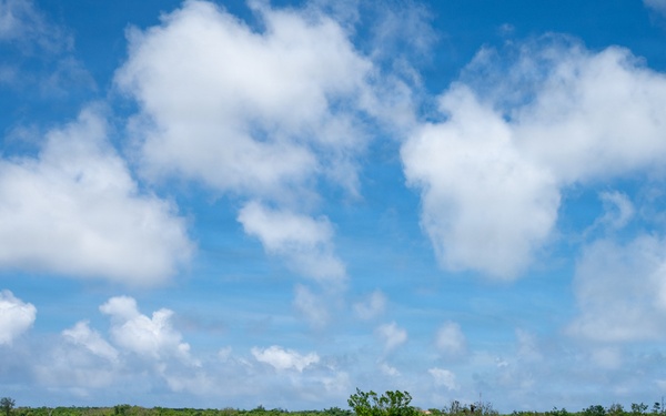 A straight two-lane road with double yellow center lines stretches toward the horizon under a partly cloudy blue sky. Military personnel and at least two camouflage-painted tactical vehicles have stopped a white civilian van on the road, with soldiers positioned on both sides of the road in what appears to be a vehicle checkpoint or interdiction exercise. The surrounding terrain consists of low tropical scrub vegetation consistent with a Pacific island environment.