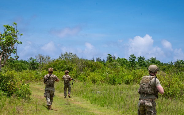 Three military personnel in full combat gear are patrolling along a grassy trail through a tropical or subtropical landscape. They are moving away from the camera in a loose file formation, carrying weapons and tactical equipment. The setting appears to be a training or operational environment with dense green vegetation under a partly cloudy blue sky.