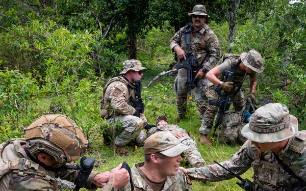 A group of military personnel in multicam/OCP camouflage uniforms are conducting what appears to be a tactical combat casualty care (TCCC) or field medical training exercise in a vegetated outdoor environment. Two soldiers are assisting a seated soldier who appears to be acting as a simulated casualty, while additional armed personnel provide security in the background. The scene is consistent with a military training exercise rather than an actual combat situation.