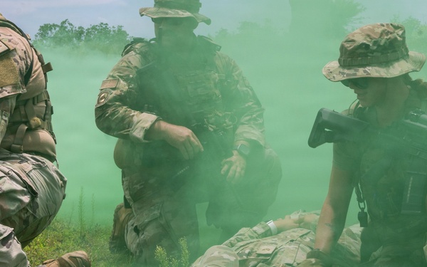 Military personnel in MultiCam/OCP camouflage uniforms are conducting a casualty evacuation (CASEVAC) or combat lifesaver training exercise in an outdoor field environment. Green smoke obscures portions of the scene, consistent with a simulated tactical scenario using smoke grenades for concealment. At least one soldier is being treated or moved on the ground while others provide security or assistance.